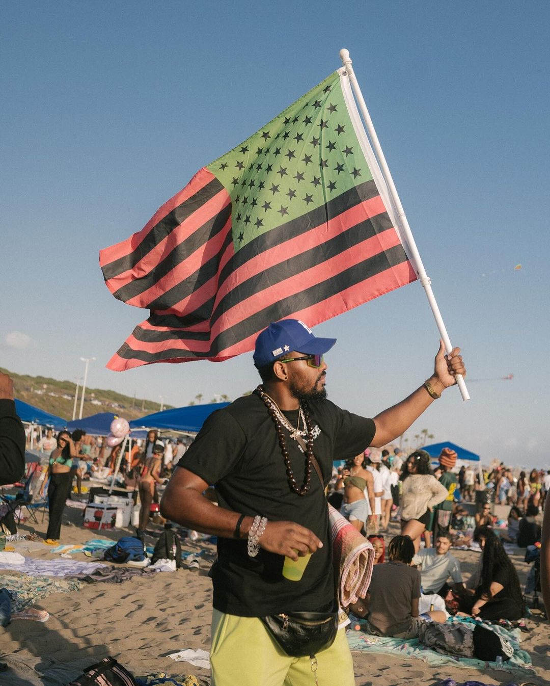 A man on a sandy beach holds a large flag with green stars and red and black stripes, photographed by Karis Beaumont in London.
