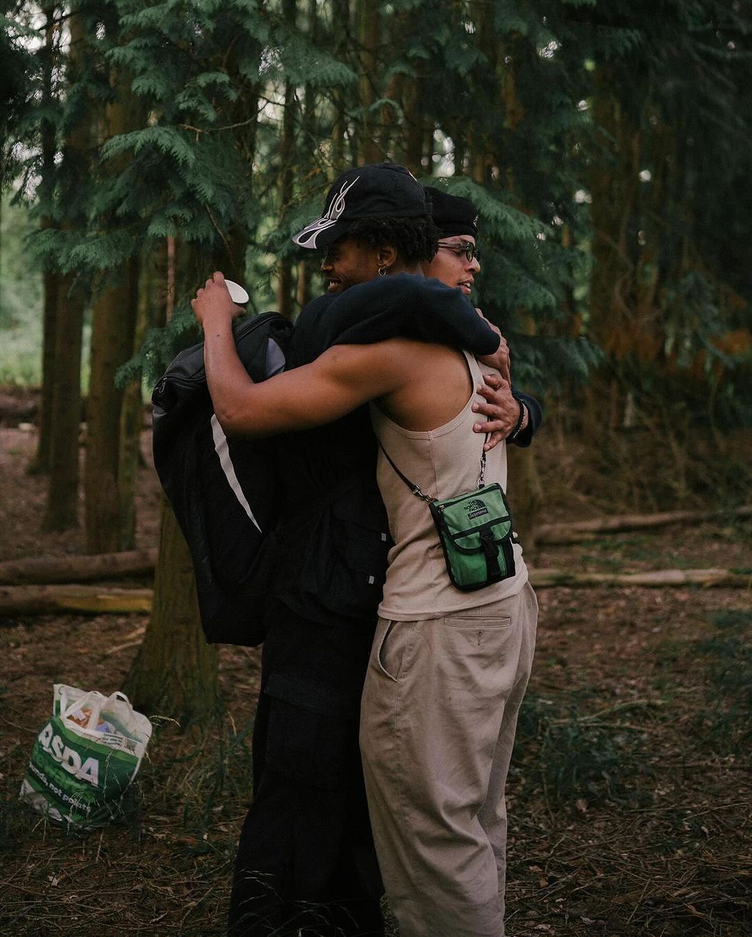 Two adult men embrace in a forest setting, surrounded by trees and foliage, with a shopping bag visible in the foreground.