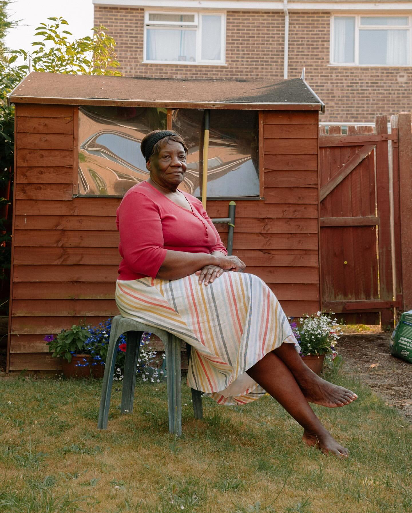 Mature Black woman in striped skirt and red shirt sits in a sunny garden on a green plastic chair in front of wooden shed.