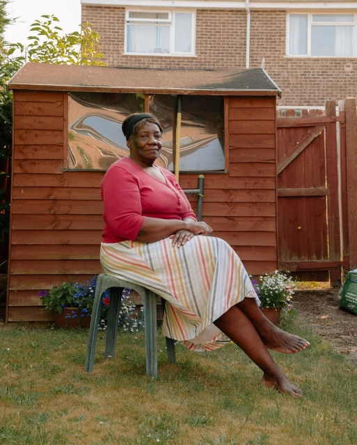 Mature Black woman in striped skirt and red shirt sits in a sunny garden on a green plastic chair in front of wooden shed.
