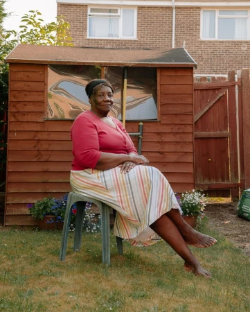 Mature Black woman in striped skirt and red shirt sits in a sunny garden on a green plastic chair in front of wooden shed.