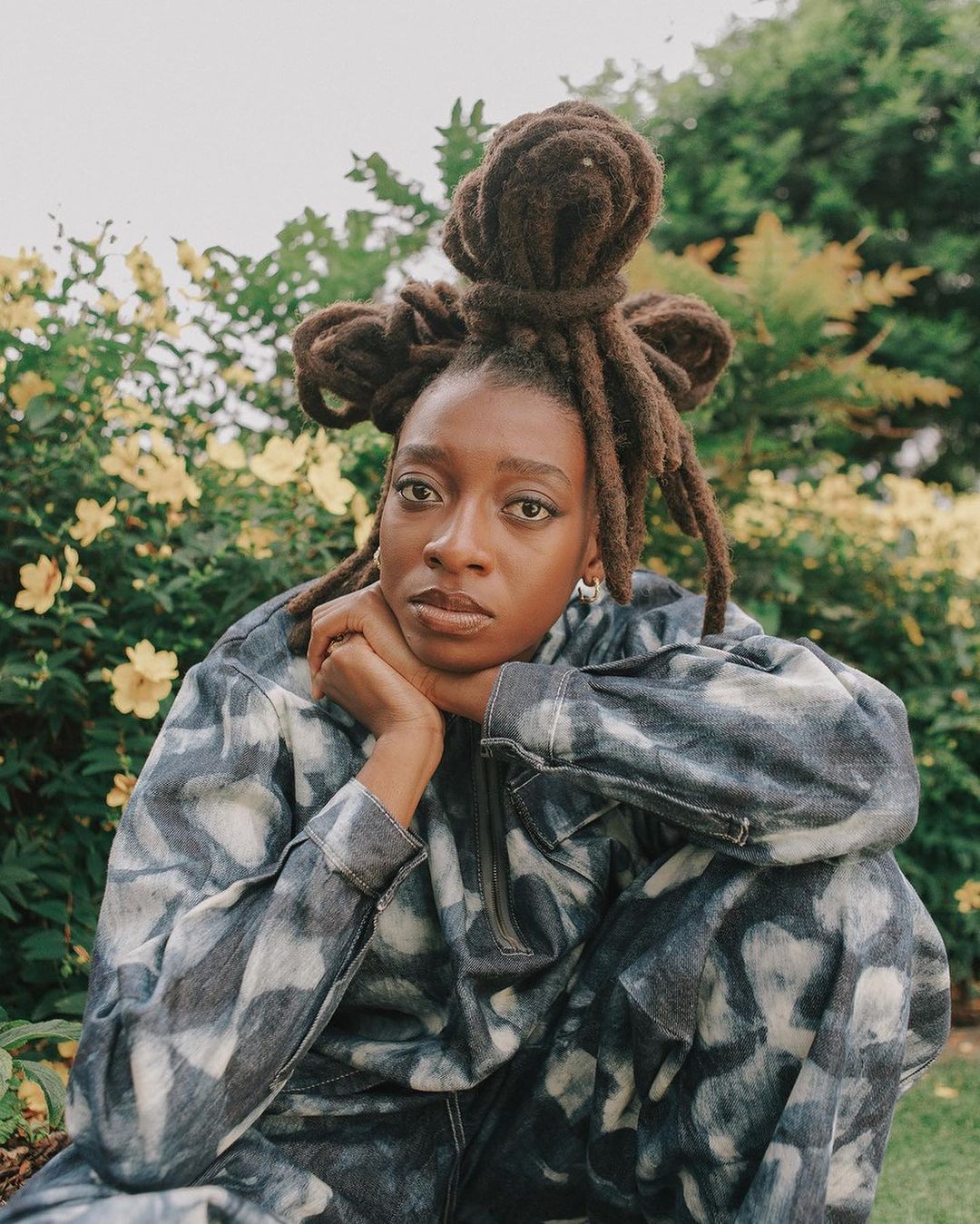 Portrait of an adult woman with dreadlocks in a patterned jumpsuit. She is sitting outside surrounded by foliage and small yellow flowers.