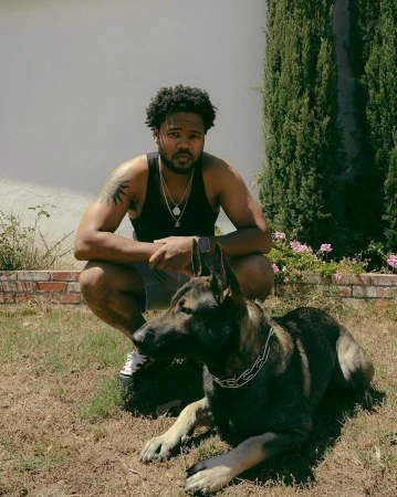 Black man crouching in a sunny yard next to a large German Shepherd dog, photographed by Karis Beaumont in London.