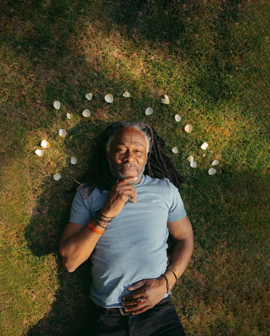 Adult man with dreadlocks lying on a grass background surrounded by white flower petals, viewed from above with a neutral expression.