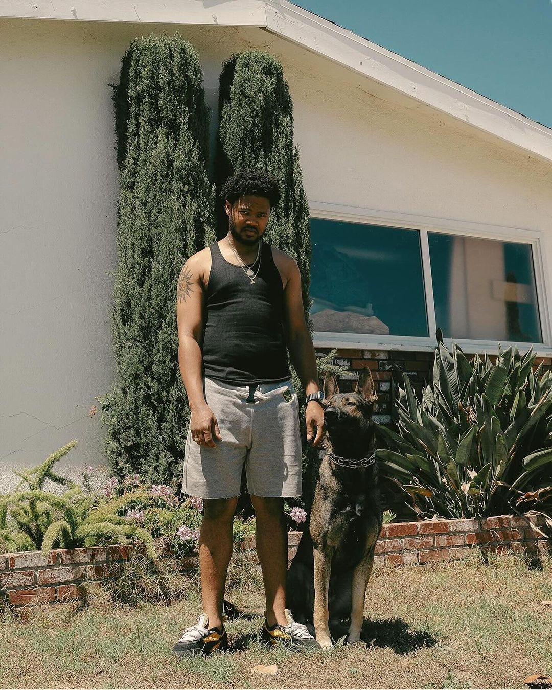 Man and dog pose outdoors near house on sunny day. Adult male in tank top stands next to German Shepherd mix dog in garden setting.