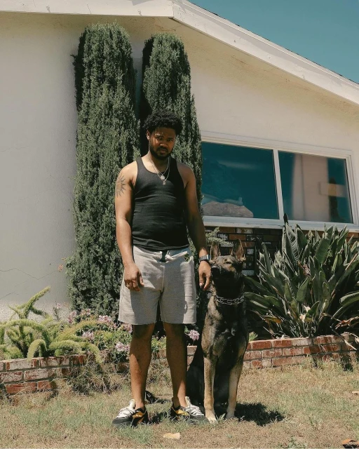 Man and dog pose outdoors near house on sunny day. Adult male in tank top stands next to German Shepherd mix dog in garden setting.