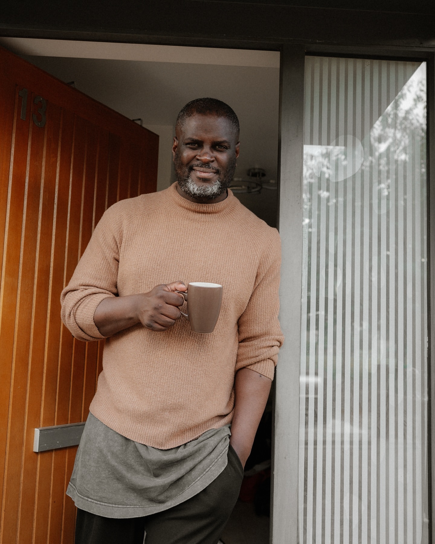 Mature adult man standing in doorway holding coffee mug. He wears a beige sweater and has a salt and pepper beard.