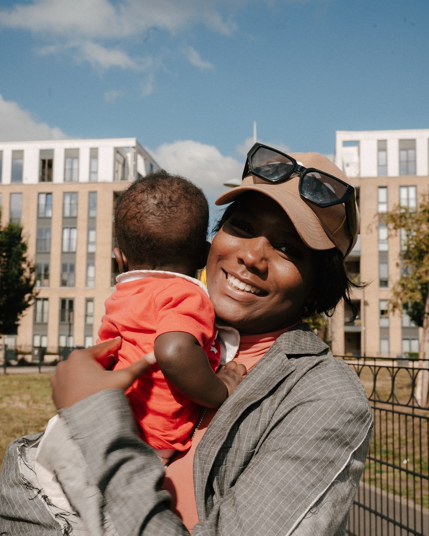 A happy African-American woman holds her baby in an outdoor urban setting with modern buildings under a sunny blue sky.