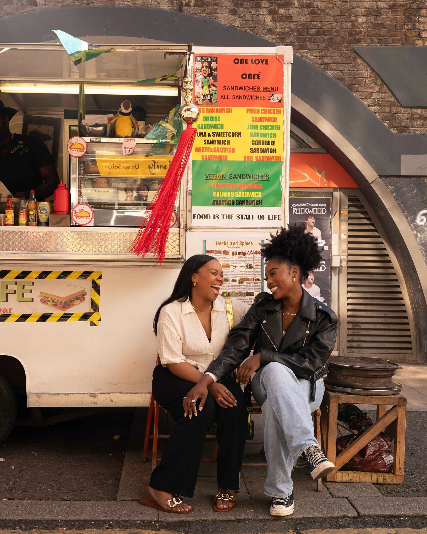 Two adult women are sitting in front of a food truck with a sandwich menu board, smiling and holding hands in an urban setting.