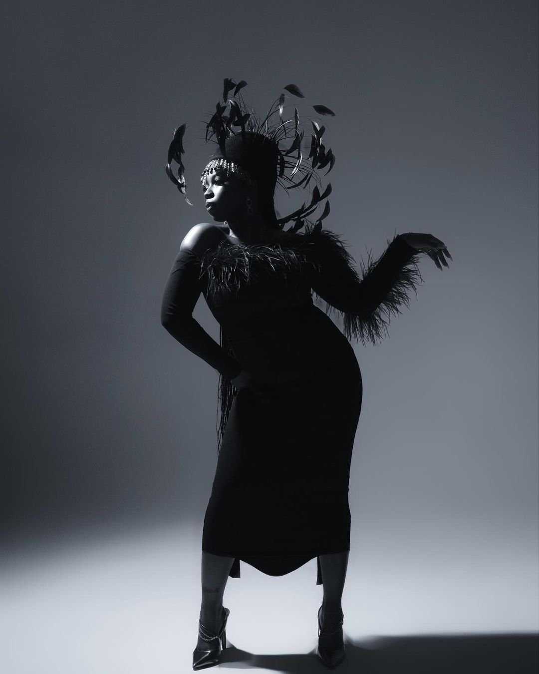 Glamorous black woman in feathered headpiece and dress striking pose against a gray backdrop in a studio photography shot.