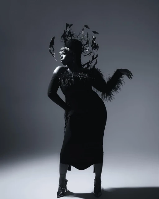 Glamorous black woman in feathered headpiece and dress striking pose against a gray backdrop in a studio photography shot.