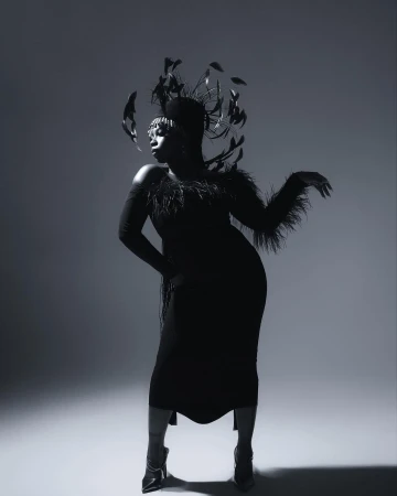 Glamorous black woman in feathered headpiece and dress striking pose against a gray backdrop in a studio photography shot.