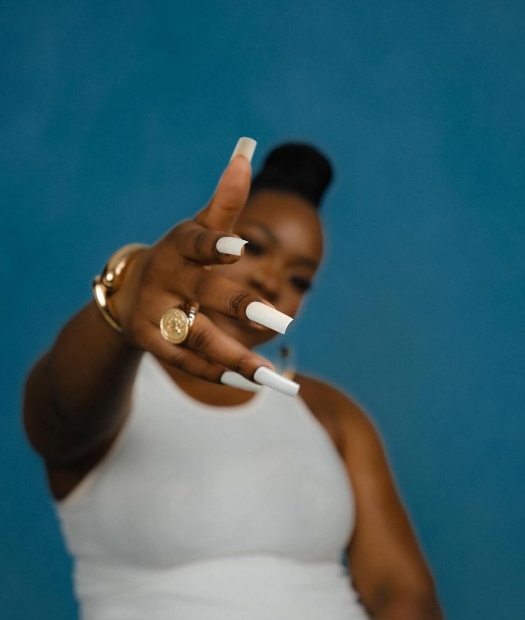 Portrait of a Black woman with long white nails, wearing gold jewelry and a white top, against a blue background, hand pointed at camera.
