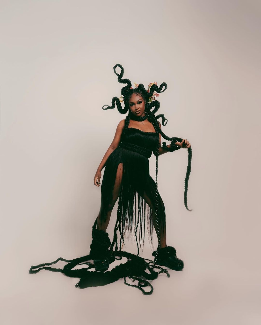 A Black female model poses in a black dress with braided hair, flowers, and chunky platform boots, against a neutral studio backdrop.