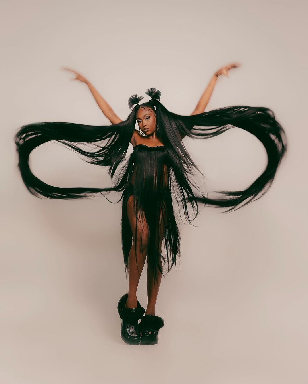 Woman with long dark hair styled in space buns and strands, wearing a strapless outfit and black platform shoes, arms raised.
