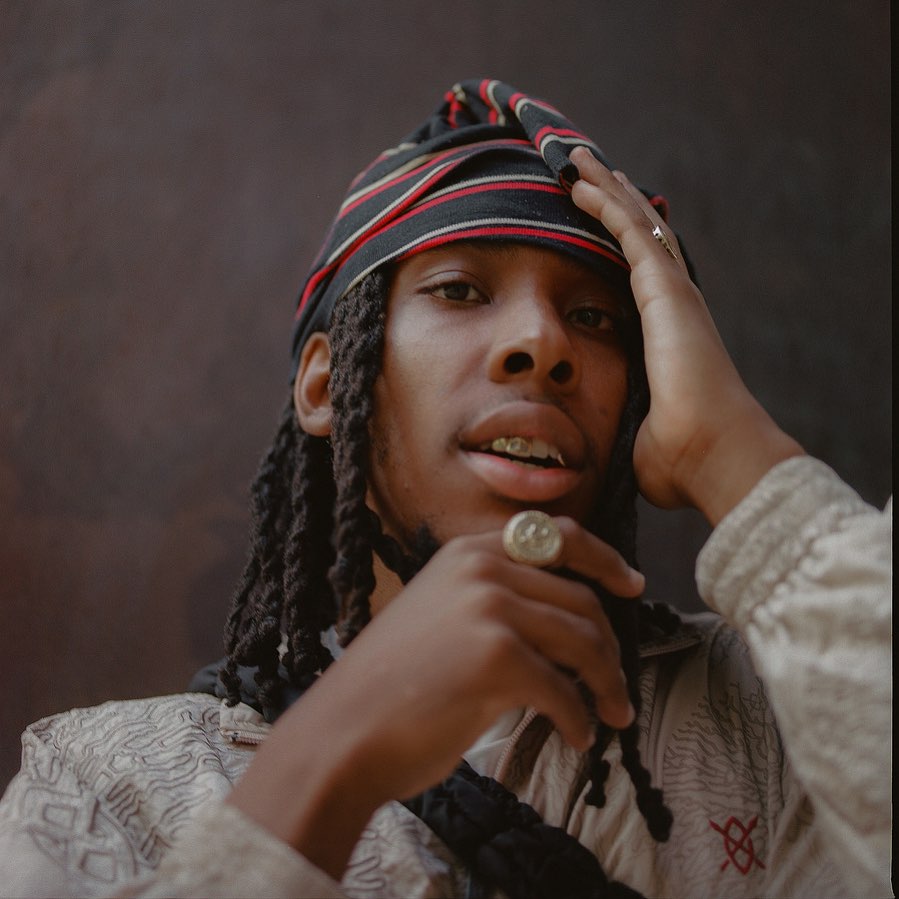 Portrait of a Black man with dreadlocks wearing a striped head covering and jewelry against a neutral brown backdrop in a photo studio.