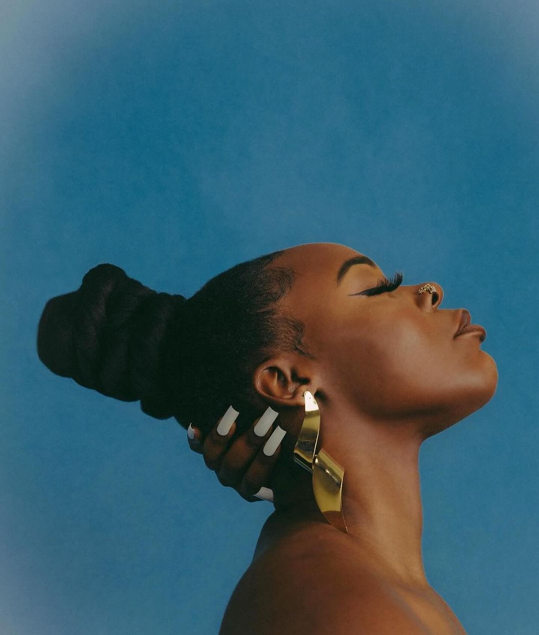 Profile of a Black woman with long eyelashes, wearing a gold earring, and her hand touching the back of her head against a blue backdrop.