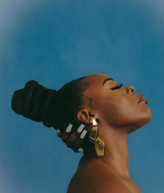 Profile of a Black woman with long eyelashes, wearing a gold earring, and her hand touching the back of her head against a blue backdrop.