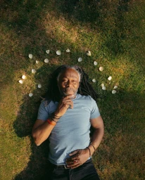An older man with long dreadlocks lying on a green lawn surrounded by a scattered circle of white petals.