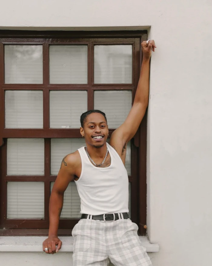 Smiling black man wearing a white tank top and plaid trousers leaning against a white wall with a brown framed window.