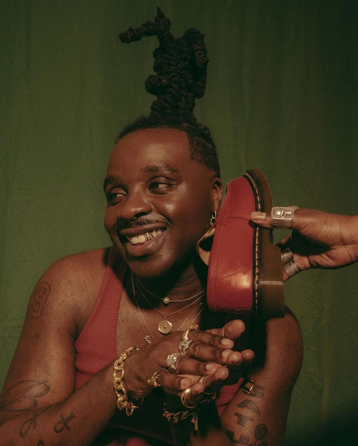Close-up portrait of a Black man with sculpted dreadlocks holding a red leather shoe near his cheek, photographed by Karis Beaumont in London.