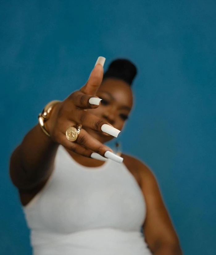 Black woman wearing a white tank top extends her hand forward, showcasing long white square nails and gold rings against a textured blue backdrop