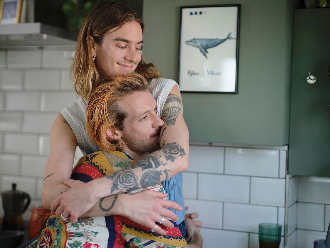 Two adult men embracing in a kitchen with natural lighting. One man has his arms around the other, with a framed whale poster in the background.