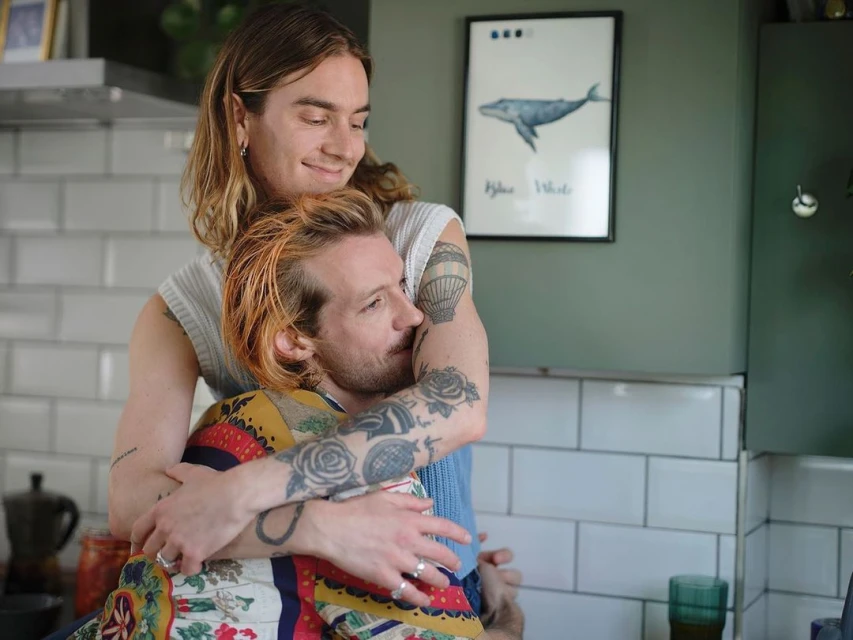 Two adult men embracing in a kitchen with natural lighting. One man has his arms around the other, with a framed whale poster in the background.