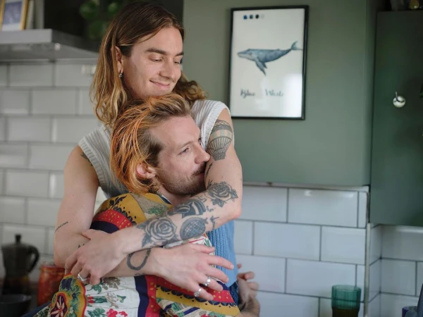 Two adult men embracing in a kitchen with natural lighting. One man has his arms around the other, with a framed whale poster in the background.