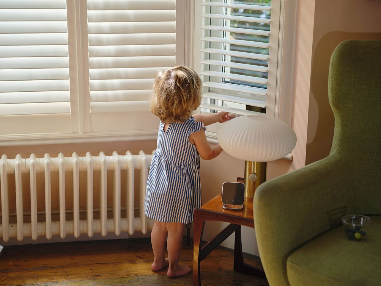 Young child in blue-and-white dress stands near window with shutters, looking at a lamp on a table, in a warmly lit room.