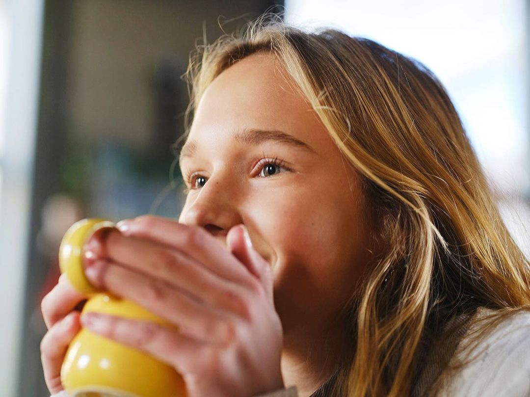 Young woman with blonde hair smiles as she holds a yellow mug indoors, enjoying a beverage, creating a cozy, warm atmosphere.