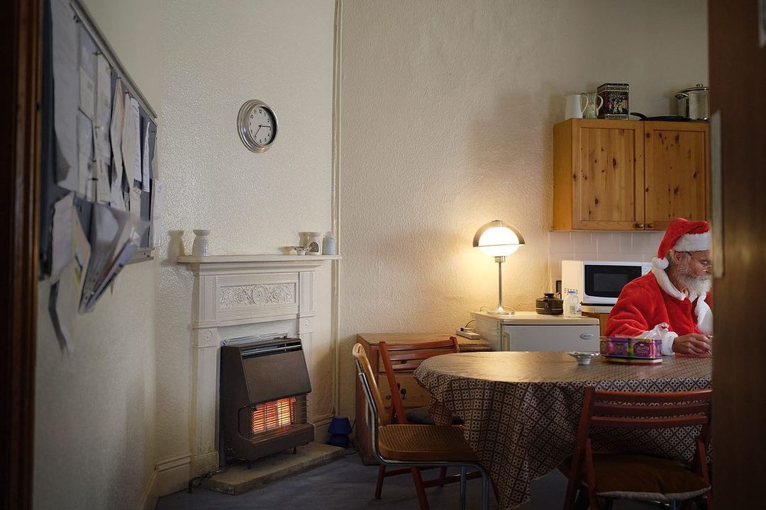 Photo of a Santa Claus character sitting at a round table in a home interior, wearing a red Santa suit and hat, next to a wall heater.