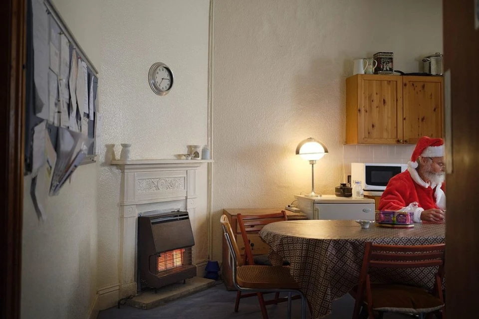 Photo of a Santa Claus character sitting at a round table in a home interior, wearing a red Santa suit and hat, next to a wall heater.