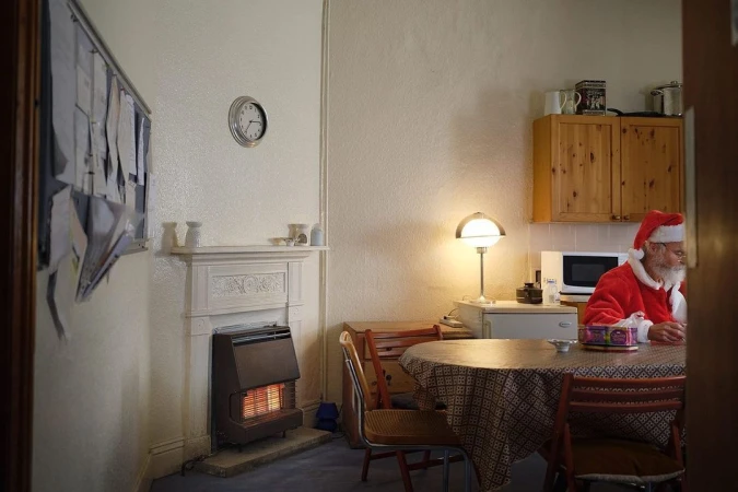 Photo of a Santa Claus character sitting at a round table in a home interior, wearing a red Santa suit and hat, next to a wall heater.
