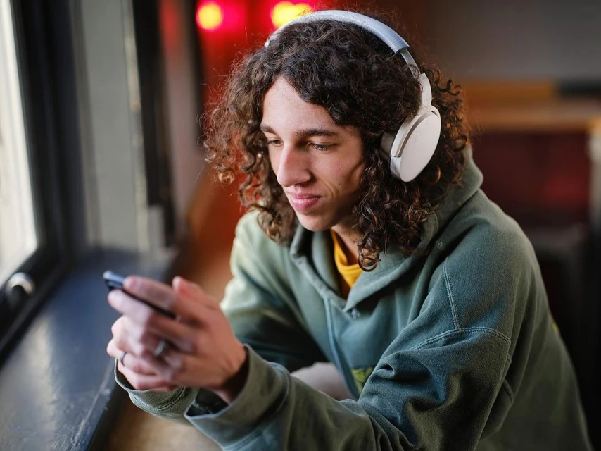Young adult male with curly hair and white headphones looks at his cell phone in a close-up shot with an interior background.