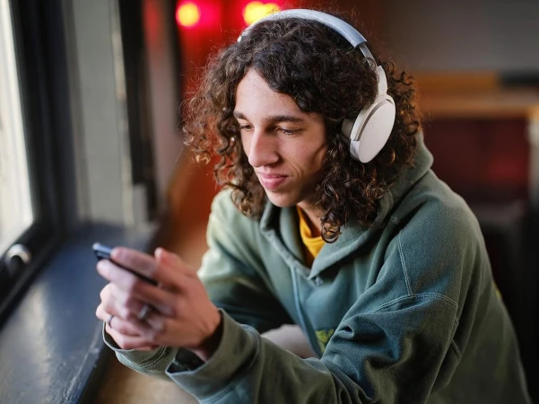 Young adult male with curly hair and white headphones looks at his cell phone in a close-up shot with an interior background.