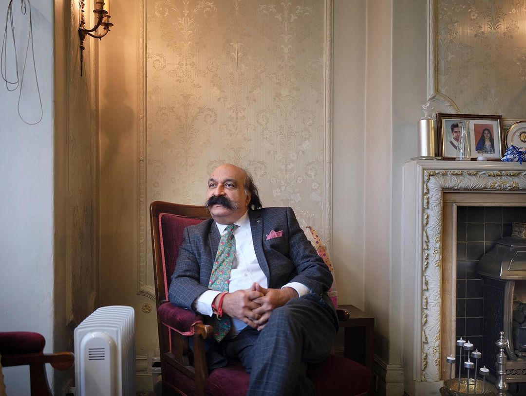 Portrait of a mature man with a mustache sitting in an armchair inside a room with patterned wallpaper and a fireplace.