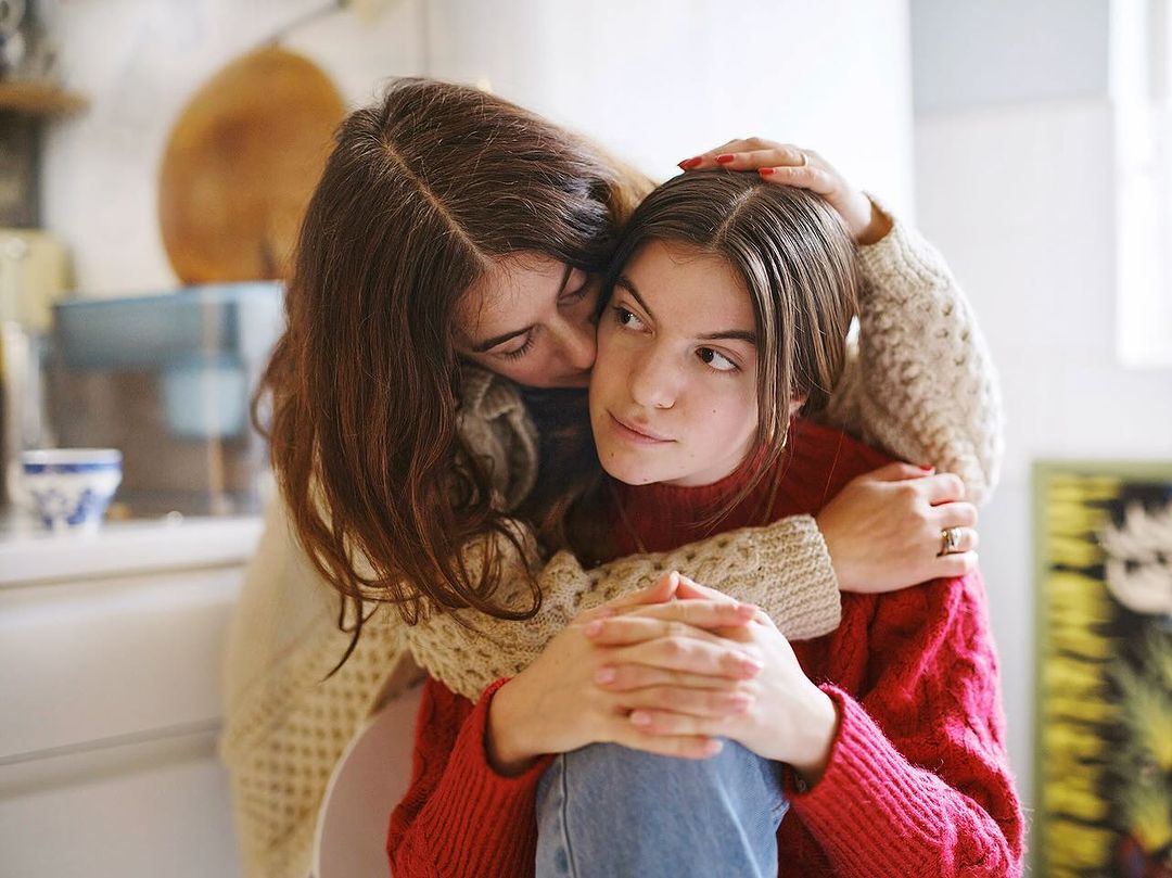 A mother embraces her teenage daughter indoors. The mother has dark hair and a cream-colored sweater. The daughter wears a red sweater.