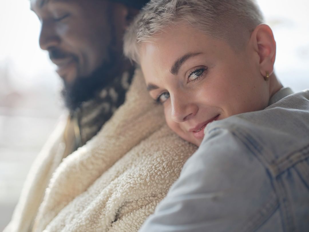 Close-up of a smiling young woman with short blond hair leaning against an African-American man in a cream-colored, sherpa-style jacket.
