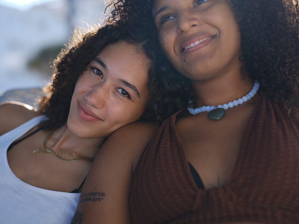 Two young women pose in an outdoor close-up. One wears a white tank top and a gold necklace, while the other wears a brown halter top.