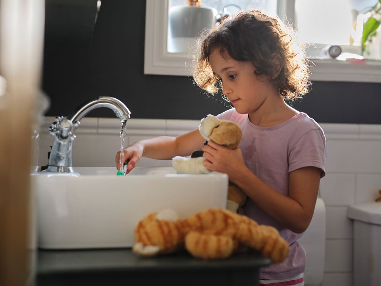 Girl washing a toothbrush at a sink while holding a plush dog and with a plush cat on the counter in a domestic bathroom.