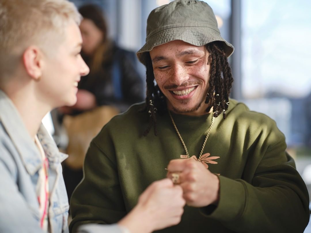 Two smiling adults touch fists indoors, one in a green sweatshirt and hat, the other in a denim jacket with bright window lighting.