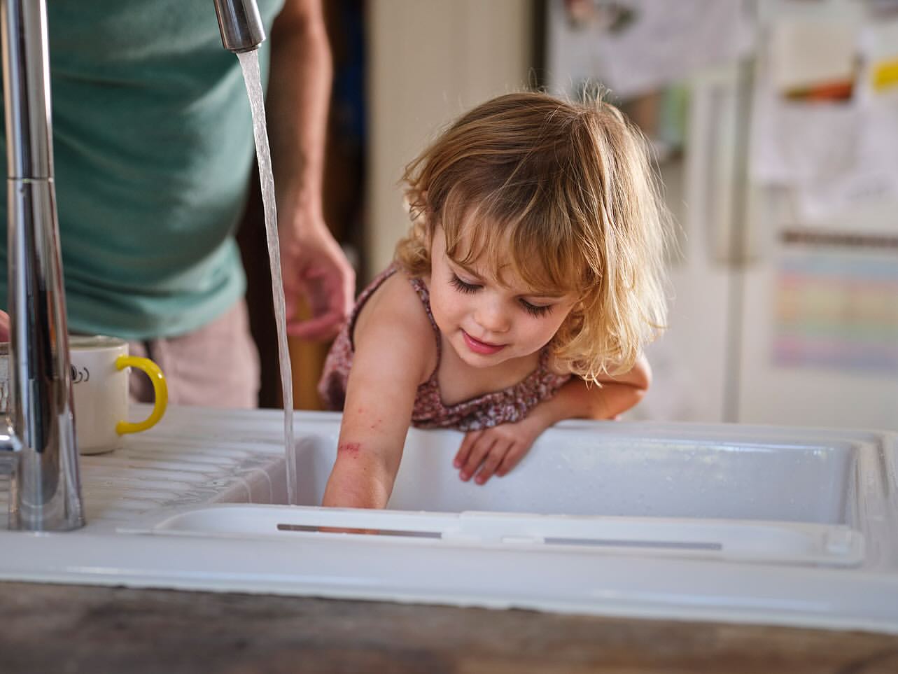 Young girl plays with running water in a white sink, a man is partially visible in the background, shot in a domestic interior.