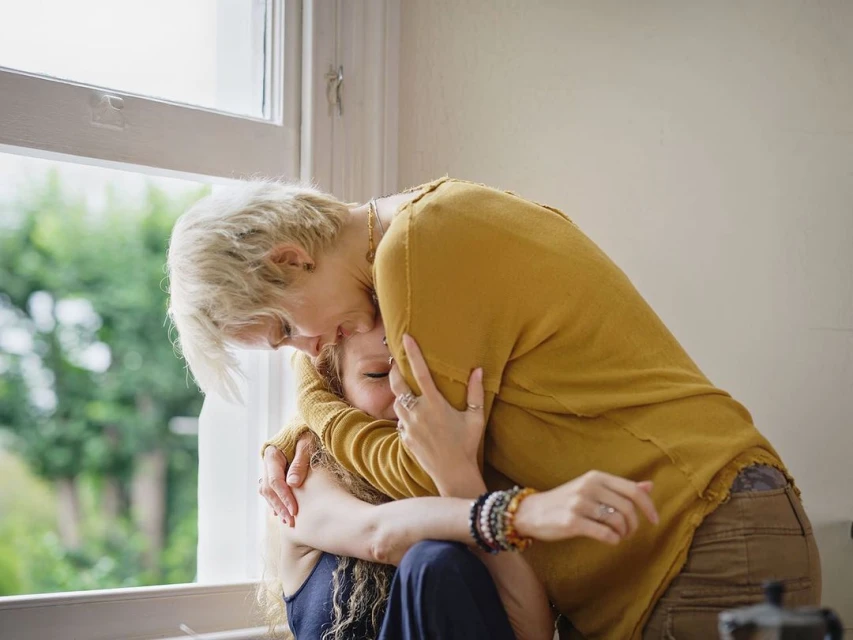 A blonde woman in yellow embraces a seated young woman near a window, creating a warm, intimate indoor scene filled with soft, natural light.