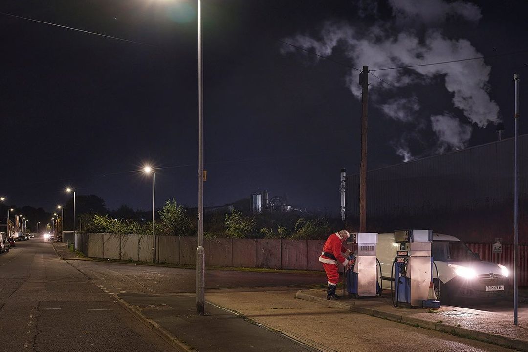 Image captures a person in a Santa Claus suit refuelling a vehicle at a gas station at night in an urban area under streetlights.