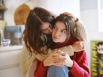 Two women embracing indoors, one wearing a cream cable-knit sweater and the other wearing a red sweater.