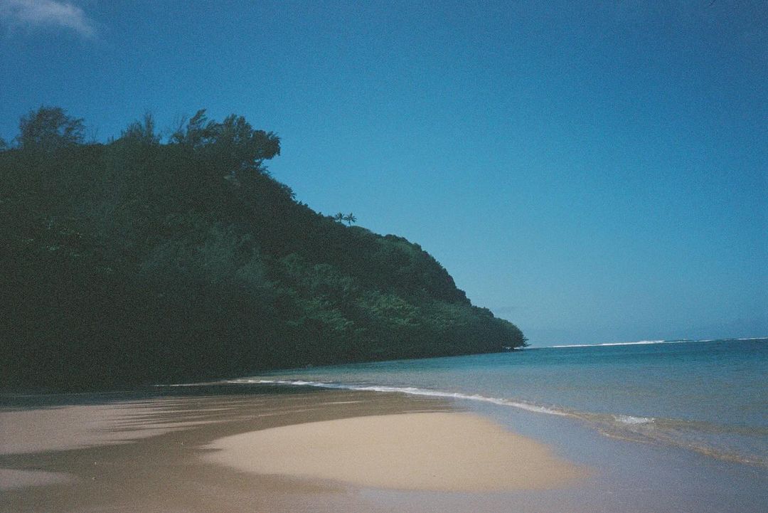 Scenic view of a tropical beach with a lush green headland, gentle waves, and clear blue sky. Serene landscape evoking relaxation and nature.