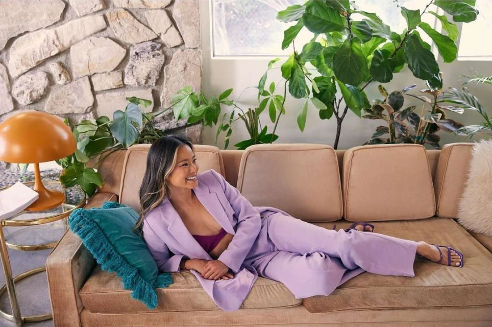 Stylish woman in a lilac suit poses on a vintage couch with green plants and a stone wall for a fashion or lifestyle shoot indoors.