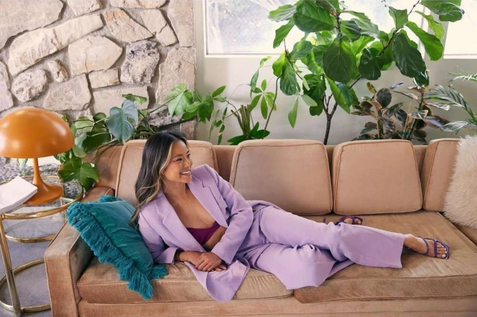 Stylish woman in a lilac suit poses on a vintage couch with green plants and a stone wall for a fashion or lifestyle shoot indoors.