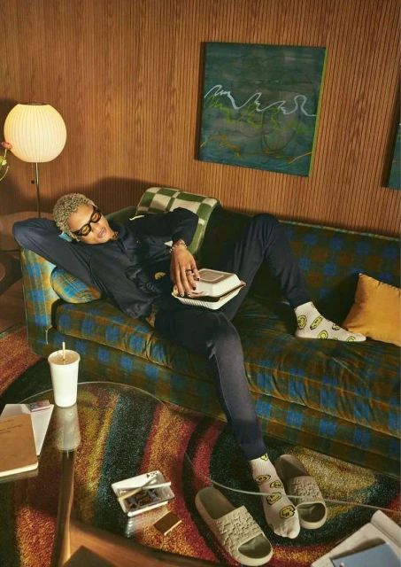Man with bleached hair lounges on a couch, wearing sunglasses, a blue jumpsuit, and socks with smiley faces in an indoor setting.
